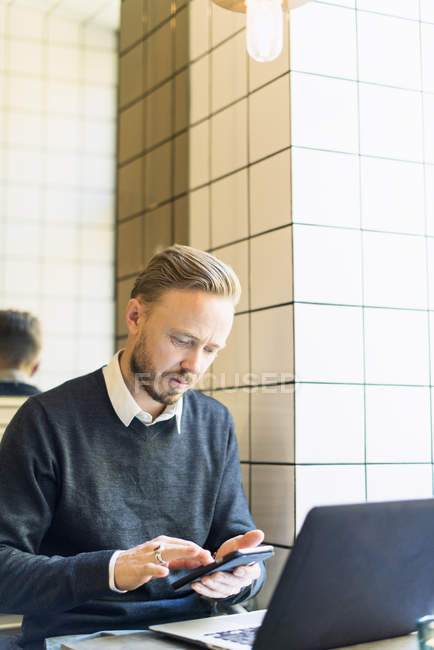 Mitte erwachsener Geschäftsmann nutzt Smartphone, während er im Café am Laptop sitzt — Stockfoto