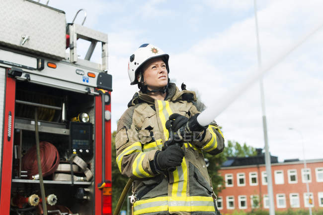 Bombeiro feminino usando mangueira de incêndio ao lado do caminhão — Fotografia de Stock