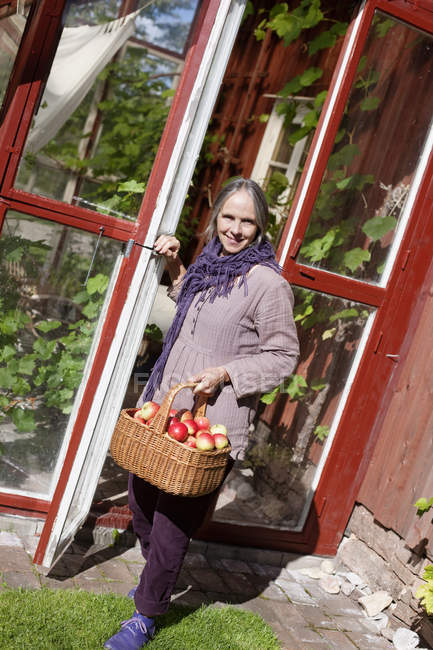 Donna anziana con cesto di mele accanto alla porta — Foto stock