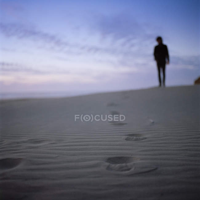 Silhouette eines Mannes, der bei Sonnenuntergang am Sandstrand steht, mit Fußabdrücken im Vordergrund — Stockfoto