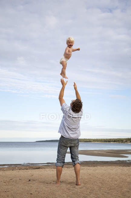 Hombre lanzando hijo en el aire en la playa contra el cielo con nubes - foto de stock