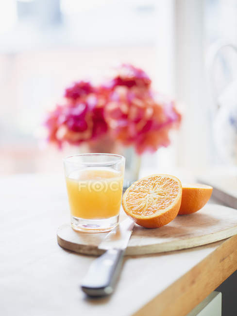 Vidro de suco de laranja com frutas cortadas pela metade na placa de corte — Fotografia de Stock
