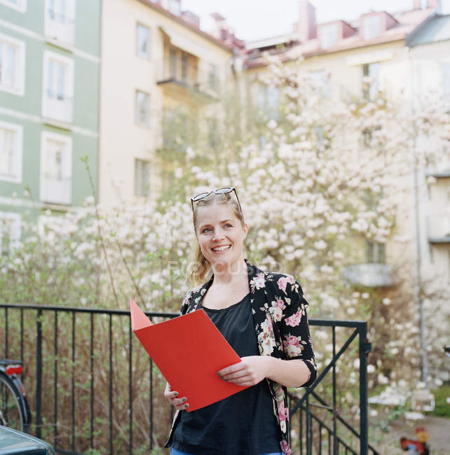 Mujer sonriente con carpeta roja en patio residencial - foto de stock