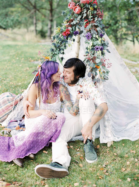 Bride and groom sitting on grass by white tent at hippie wedding — Stock Photo