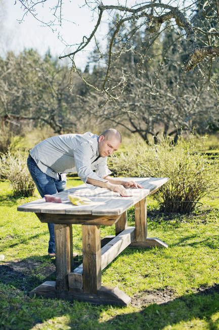 Homem adulto médio polimento mesa de madeira no jardim — Fotografia de Stock