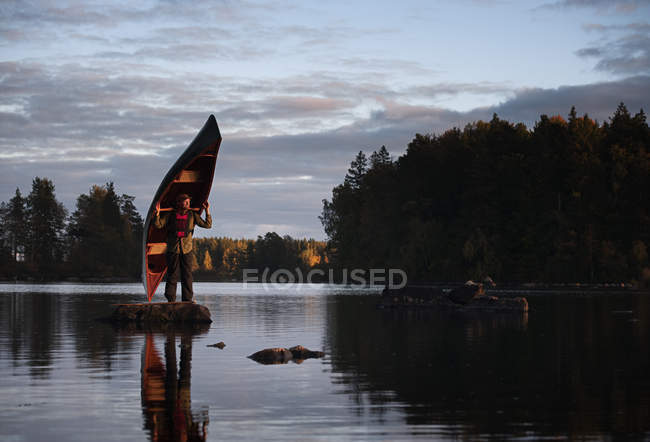 Hombre de pie sobre piedra en el lago sosteniendo canoa - foto de stock