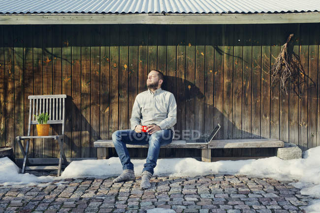 Hombre adulto sentado en el banco junto a un cobertizo de madera - foto de stock