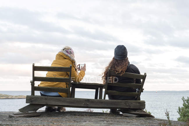 Вид сзади на молодых женщин, сидящих на деревянных стульях у озера и фотографирующих — стоковое фото
