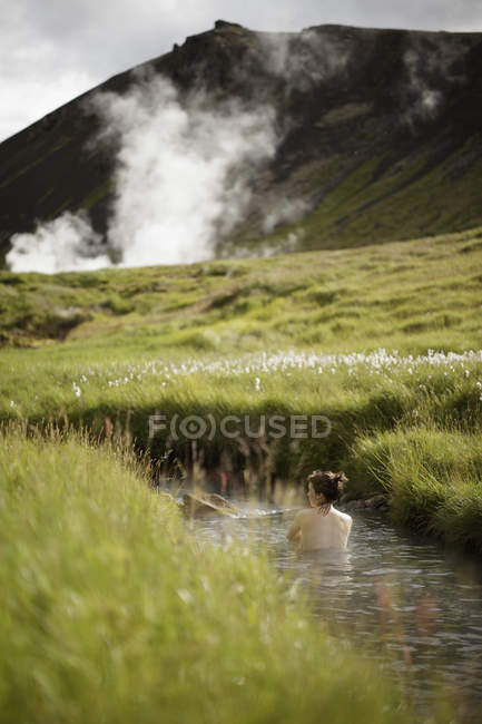 Задний вид женщины, купающейся в потоке в Исландии с гейзером и горой на заднем плане — стоковое фото