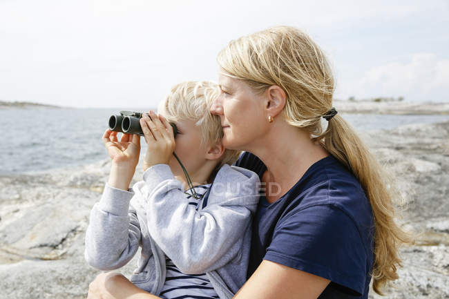 Mãe e filho sentados na costa rochosa, filho olhando através de binóculos no arquipélago de Estocolmo — Fotografia de Stock