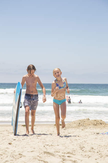 Niño con tabla de surf y niña caminando en la playa en San Diego - foto de stock