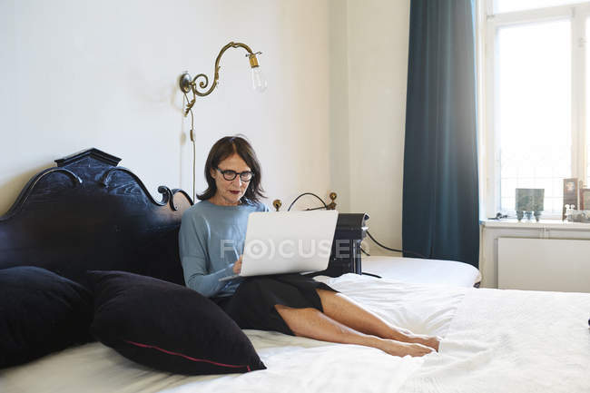 Доросла жінка використовує ноутбук, сидячи на ліжку — стокове фото