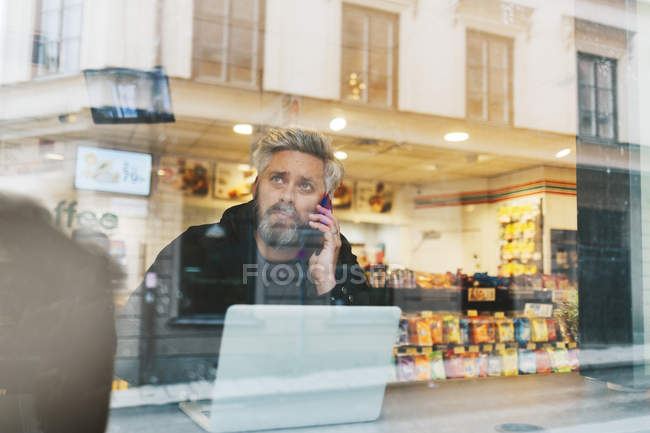 Man talking on smart phone by laptop behind window — Stock Photo