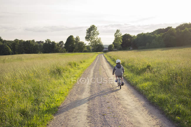 Boy ciclismo a lo largo de la carretera terrestre que conduce a través de campos - foto de stock