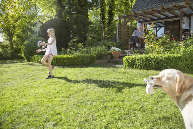Chica jugando con el perro en frente de la casa, hombre sentado en el fondo - foto de stock