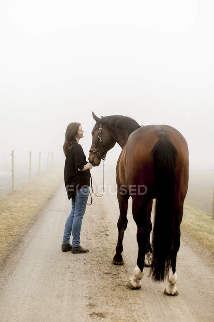 Mujer adulta con caballo en el camino de tierra en la niebla - foto de stock