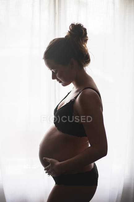 Vista lateral da mulher grávida em pé pela janela — Fotografia de Stock