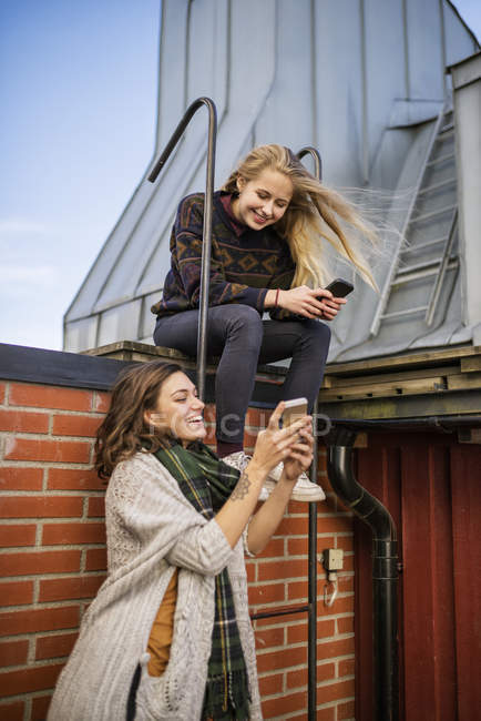 Dos mujeres jóvenes usando teléfonos en el techo, enfoque selectivo - foto de stock