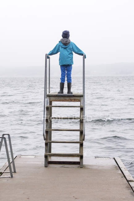Visão traseira do menino em pé na torre de mergulho construída na borda do cais — Fotografia de Stock