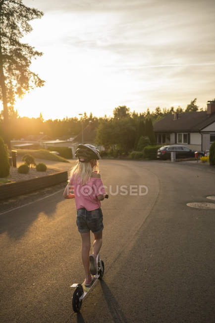 Rear view of girl riding push scooter along town street, focus on foreground — Stock Photo