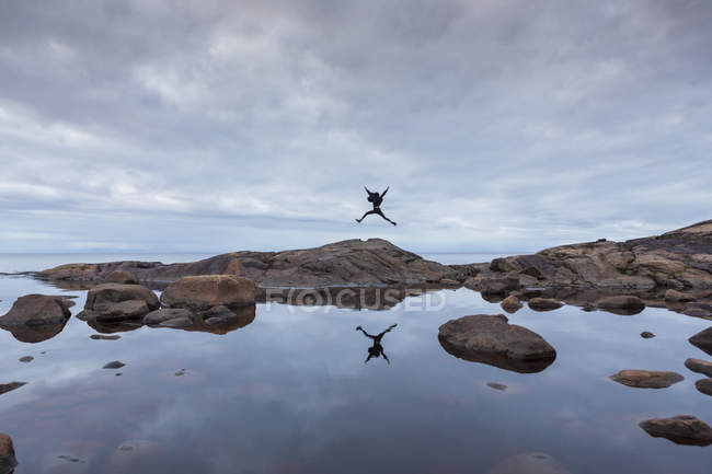 Vista trasera de la mujer saltando sobre rocas por lago bajo cielo nublado - foto de stock