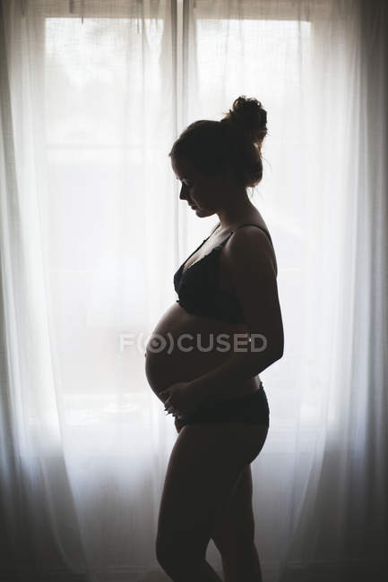 Vista lateral de la mujer embarazada de pie por la ventana - foto de stock