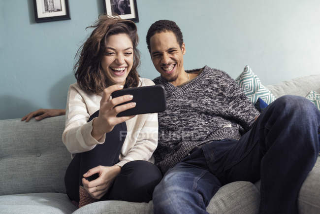 Молода пара сидить на дивані, дивлячись на смартфон і сміючись — стокове фото