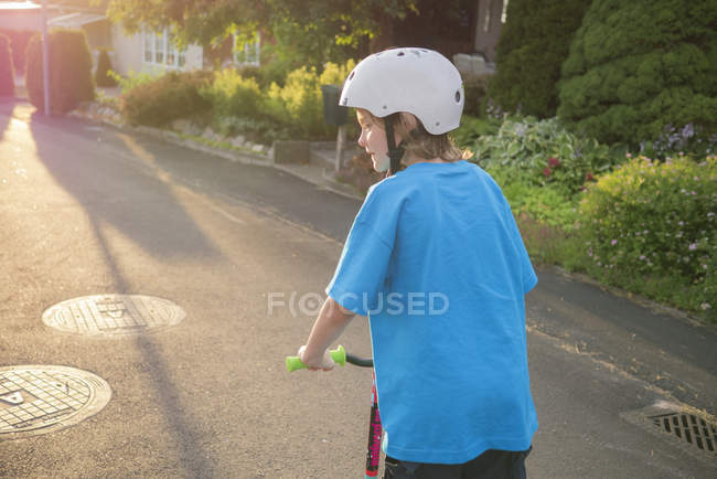 Visão traseira do menino montando empurrar scooter ao longo da rua da cidade — Fotografia de Stock