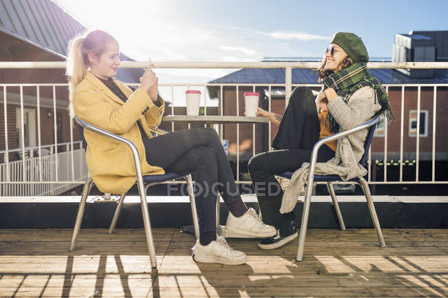Дві молоді жінки, використовуючи смартфон, сидять за столом і п'ють каву — стокове фото