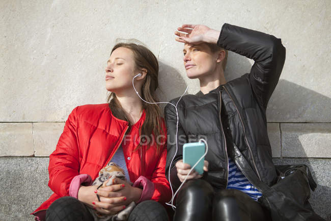 Dos mujeres escuchando música en el teléfono inteligente mientras están sentadas en los escalones - foto de stock