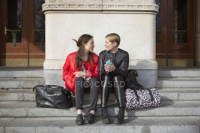 Two women listening music on smartphone while sitting on steps — Stock Photo