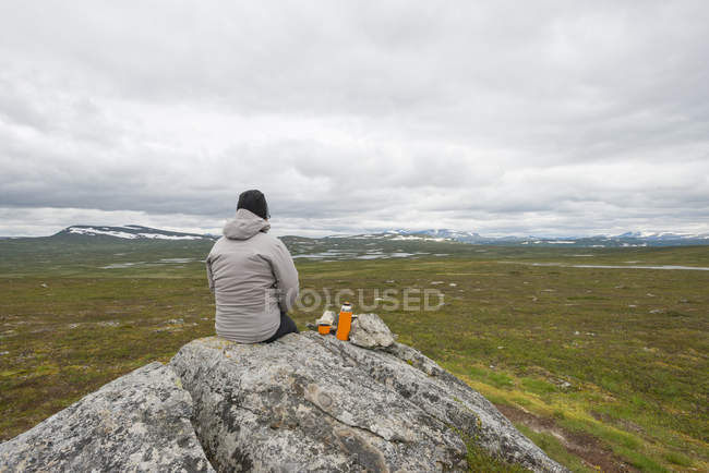 Femme avec récipient à boisson isolé assis sur la roche et regardant les montagnes — Photo de stock