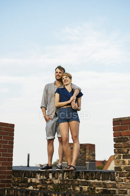 Улыбающаяся молодая пара обнимается, стоя на крыше — стоковое фото