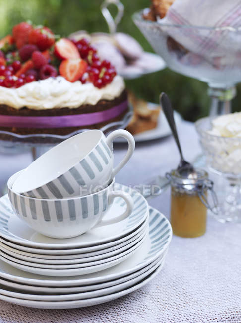 Tazas de té y platos en la mesa con pastel en el fondo, se centran en primer plano - foto de stock