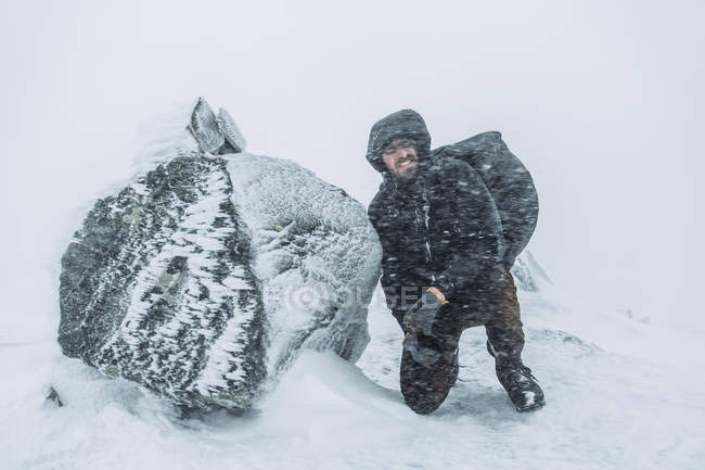 Молодой турист, стоящий на коленях рядом со скалой во время снежной метели — стоковое фото