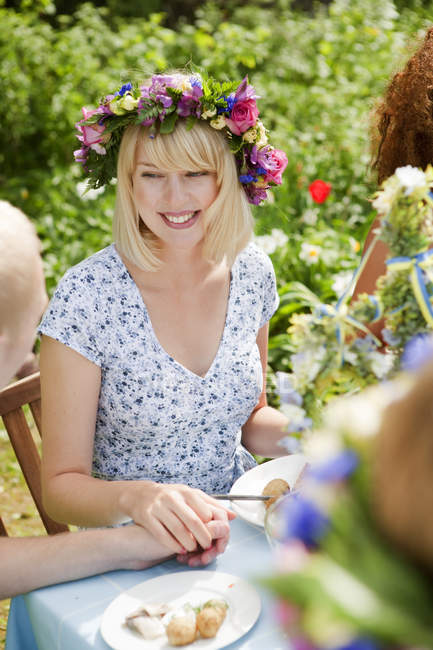 Mujer joven con corona de flores sentada en la mesa de picnic y sosteniendo la mano masculina - foto de stock