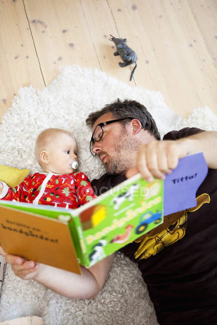 Vista elevada de padre e hija tumbados en el suelo y leyendo libro - foto de stock