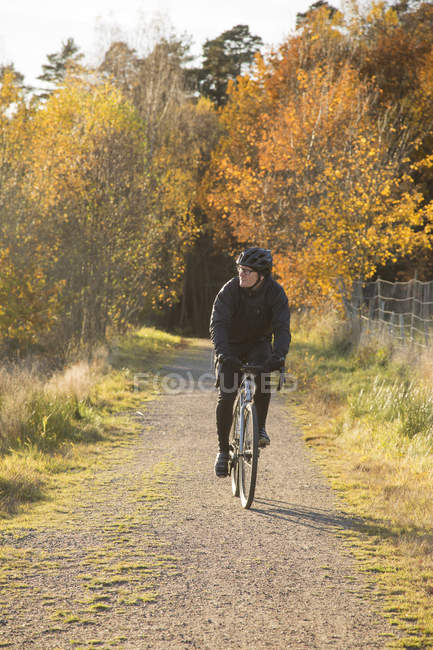 Mature man riding bicycle on dirt road through autumn forest — Stock Photo