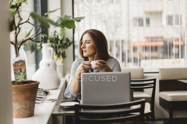 Mujer sosteniendo la taza con café por ordenador portátil en la cafetería - foto de stock