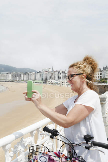 Mid adult woman taking photo on smart phone on beach in San Sebastian, Spain — Stock Photo