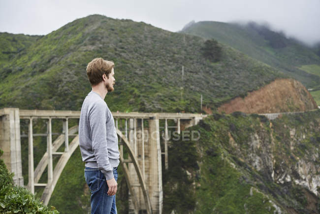 Man standing in front of bridge and mountains at Big Sur in California, USA — Stock Photo