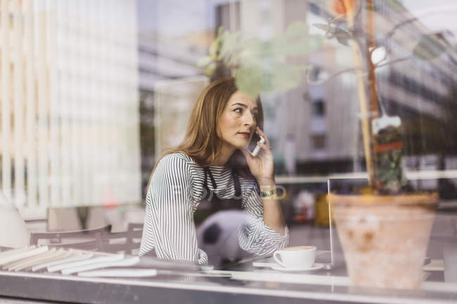 Mujer usando el teléfono inteligente detrás de la ventana de la cafetería, enfoque selectivo - foto de stock