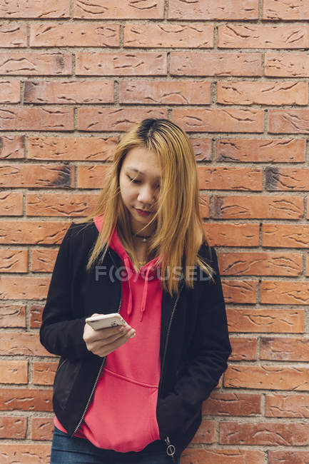 Девочка-подросток, прислонившись к кирпичной стене, смотрит на мобильный телефон — стоковое фото