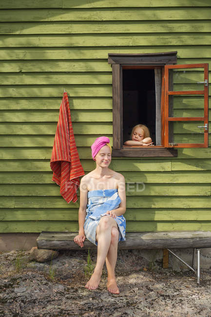 Mujer adulta sentada fuera de la sauna, hija mirando por la ventana - foto de stock