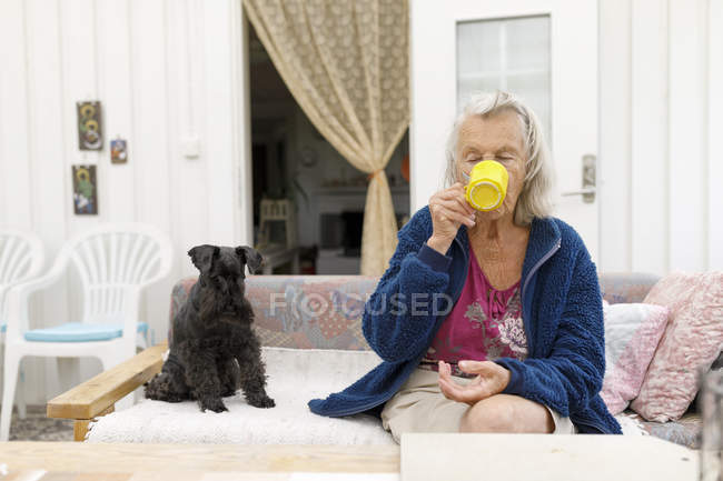 Mulher com cão no sofá beber de copo amarelo, foco em primeiro plano — Fotografia de Stock