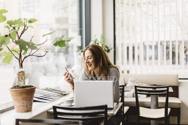 Mulher usando telefone inteligente por laptop no café, foco seletivo — Fotografia de Stock