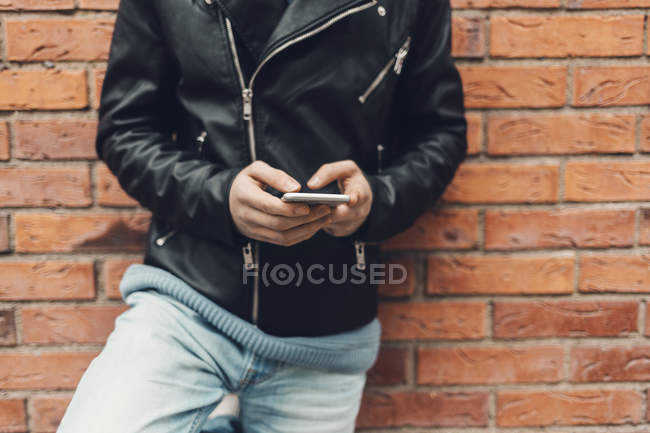 Vista recortada del adolescente apoyado en la pared de ladrillo, utilizando el teléfono inteligente, enfoque selectivo - foto de stock