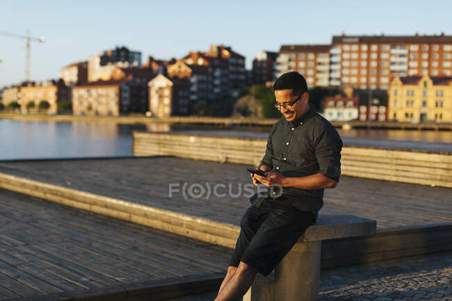 Hombre sentado en el paseo marítimo utilizando teléfonos inteligentes y sonriendo al sol. - foto de stock