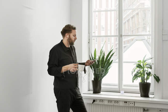 Homme dans les écouteurs utilisant un smartphone et se tenant debout devant une fenêtre — Photo de stock