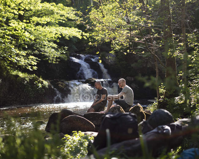 Hombres sentados en rocas por cascada, enfoque selectivo - foto de stock